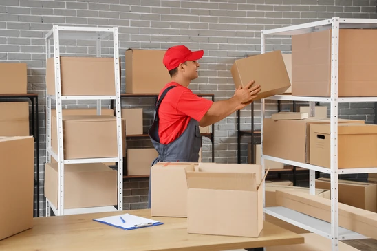 Livreur en uniforme tenant un colis cartonn&eacute; dans un entrep&ocirc;t logistique avec pile de cartons d'exp&eacute;dition